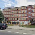 Front view of a multi-story training institute building with a beige and red facade and a sign reading 'Zakład Doskonalenia Zawodowego' atop the roof, surrounded by a brick fence and parked cars.