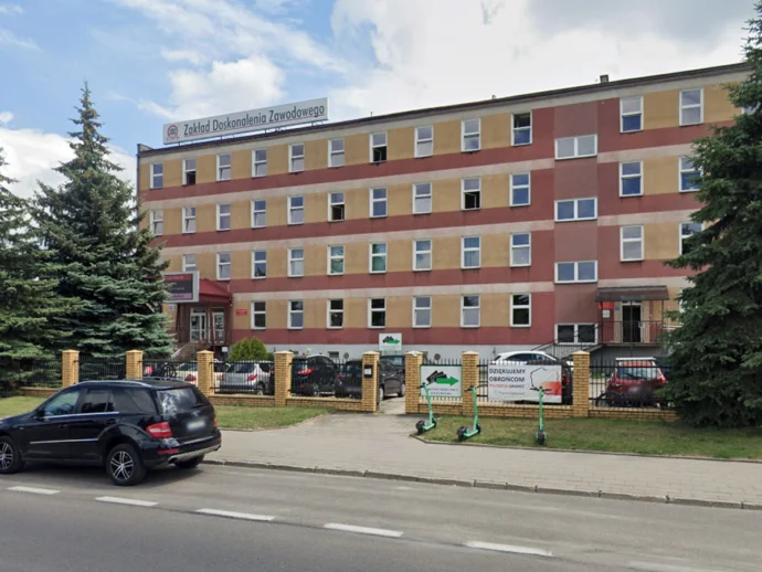 Front view of a multi-story training institute building with a beige and red facade and a sign reading 'Zakład Doskonalenia Zawodowego' atop the roof, surrounded by a brick fence and parked cars.