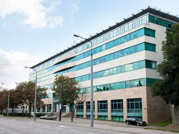 Modern glass-front office building along a street with trees and parked cars on a sunny day.