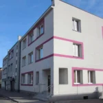 White multi-story apartment building with pink rectangular trim along windows, on a sunny street.