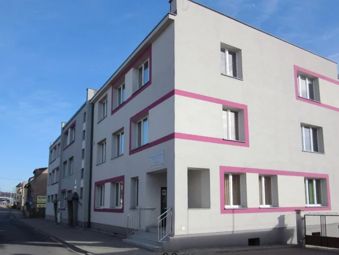 White multi-story apartment building with pink rectangular trim along windows, on a sunny street.