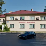 Two-story beige building with a red-tiled roof and a red sign reading 'Powiatowy Urząd Pracy' on the front, a black car parked on the street, and trees nearby.