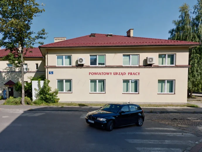 Two-story beige building with a red-tiled roof and a red sign reading 'Powiatowy Urząd Pracy' on the front, a black car parked on the street, and trees nearby.