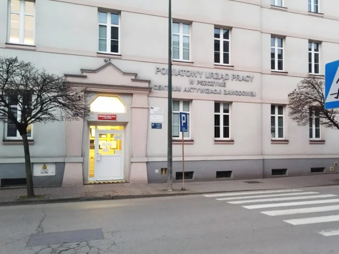 Building housing a job center with a white entrance door lit from inside and a pale pink facade, Polish signage above the door and a crosswalk in front