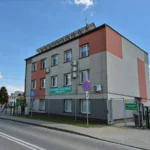 Three-story public office building with grey and pink panels on a sunny street; sign reads Powiatowy Urząd Pracy w Myślenicach (District Employment Office).