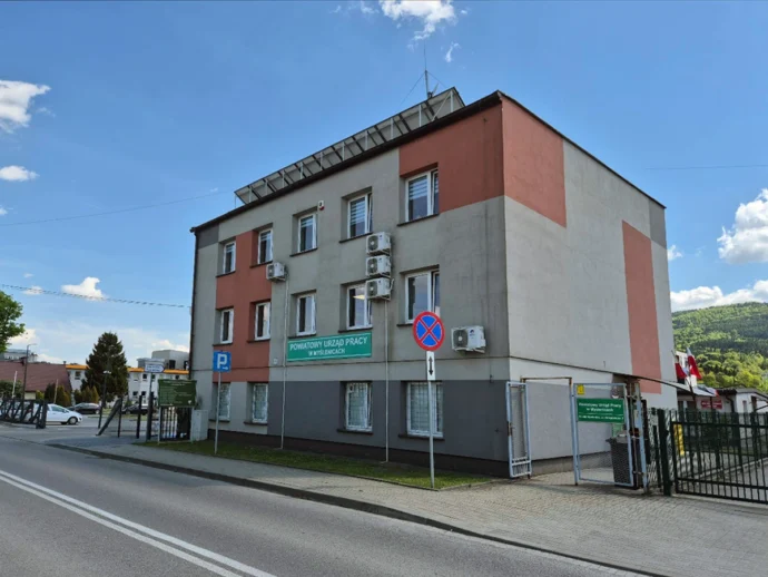 Three-story public office building with grey and pink panels on a sunny street; sign reads Powiatowy Urząd Pracy w Myślenicach (District Employment Office).