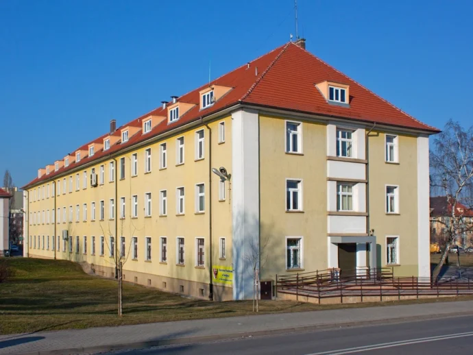 Three-story yellow apartment building with a red gabled roof along a street under a clear blue sky.