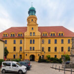 Yellow historic building with a tall green-topped tower, red-tiled roof, and arched windows in a courtyard setting with parked cars and a stone fountain on the right.