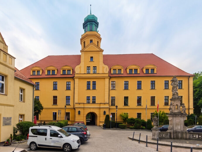 Yellow historic building with a tall green-topped tower, red-tiled roof, and arched windows in a courtyard setting with parked cars and a stone fountain on the right.