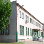 Two-story pale pink building with green window bars, a tree on the left, and a bicycle near the entrance by a fence lline.