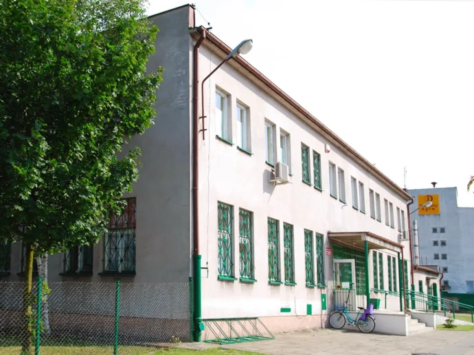 Two-story pale pink building with green window bars, a tree on the left, and a bicycle near the entrance by a fence lline.