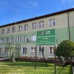 Three-story light-green government office building with a blue sky, glass doors, and a prominent green sign at the front reading Powiatowy Urząd Pracy w Gostyninie.