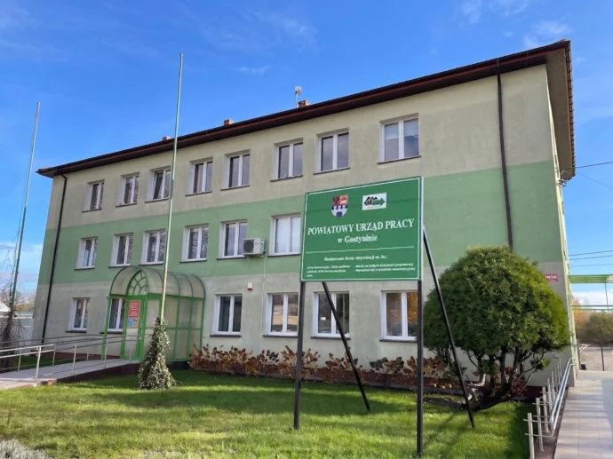 Three-story light-green government office building with a blue sky, glass doors, and a prominent green sign at the front reading Powiatowy Urząd Pracy w Gostyninie.