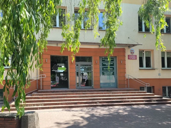 Entrance to a public building with glass doors, brick steps, and overhanging green tree branches partially obscuring the view.