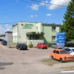 Green government office building with a parking lot and blue auto shop sign in Polish reading INTER AUTO and parts store nearby car park visible in front.