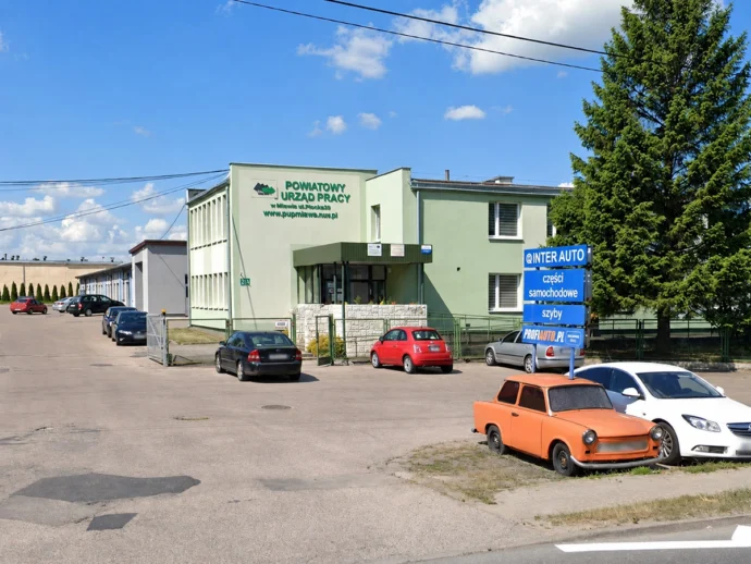 Green government office building with a parking lot and blue auto shop sign in Polish reading INTER AUTO and parts store nearby car park visible in front.