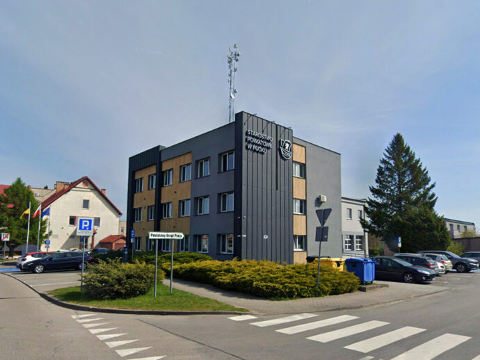 Modern municipal building with signage and a parking lot in front; blue municipal bins nearby and a crosswalk in foreground