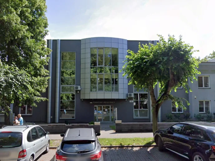 Front view of a modern office building with a curved glass facade and a sign reading 'Powiatowy Urząd Pracy' above the entrance, trees and parked cars in front.