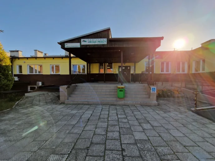 Front of a public employment office (Urząd Pracy) with a yellow and brown exterior, a dark canopy over the entrance, stairs and a ramp leading to the doors, sunny day with sun flare to the right.