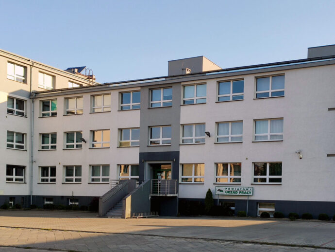 White multi-story office building with a central dark entrance and a sign reading "Powiatowy Urząd Pracy" by the ground-floor windows.