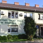 Pale green two-story building with a sign reading 'POWIATOWY URZĄD PRACY W ŻUROMINIE' and several windows, surrounded by bushes and a small parking area sign in front.