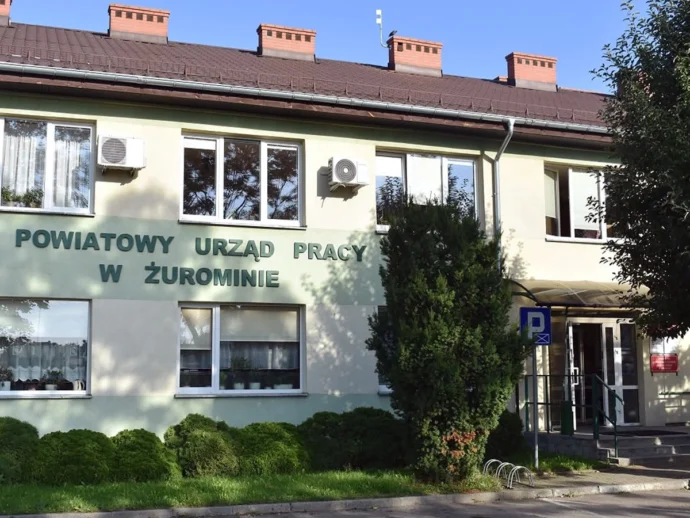 Pale green two-story building with a sign reading 'POWIATOWY URZĄD PRACY W ŻUROMINIE' and several windows, surrounded by bushes and a small parking area sign in front.