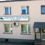 Facade of Powiatowy Urząd Pracy w Rykach, a beige, two‑story building with a large sign above the ground floor windows and an entrance on the right.
