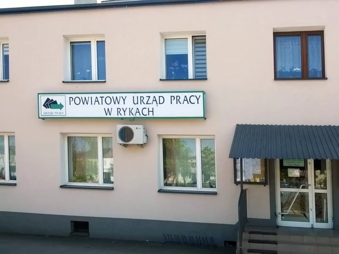 Facade of Powiatowy Urząd Pracy w Rykach, a beige, two‑story building with a large sign above the ground floor windows and an entrance on the right.