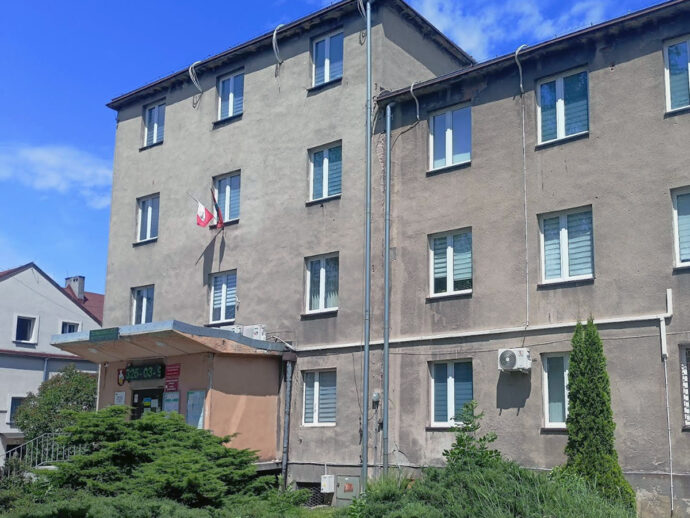 Three-story beige apartment building with a small entrance canopy and flags hanging from the wall, blue sky above.