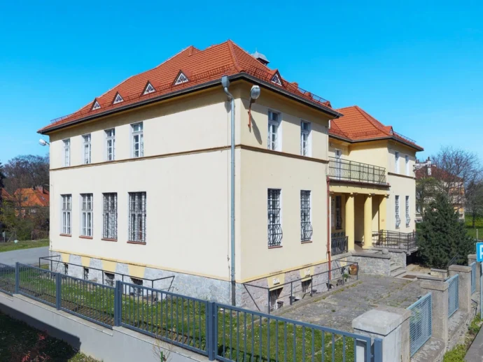 Two-story beige building with a red tiled roof, decorative wrought-iron window bars, and a small front entrance porch with columns.