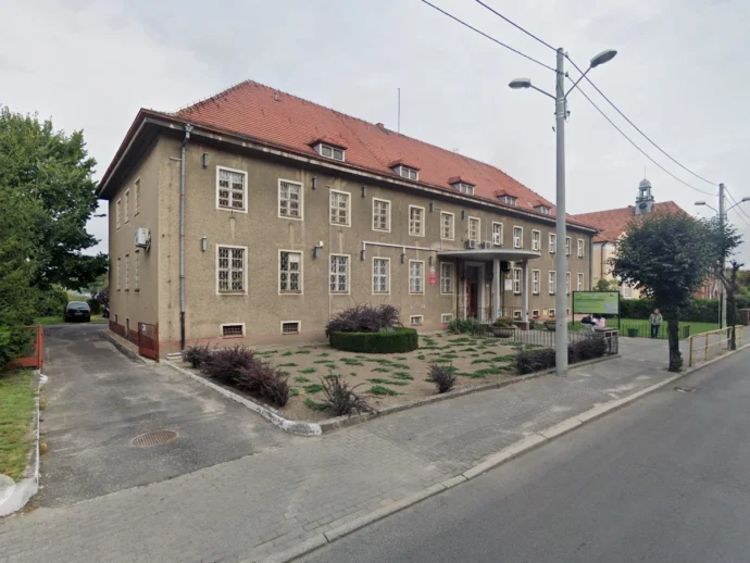 Two-story beige building with a red tile roof and small landscaped front yard, situated along a paved street with a sidewalk and streetlights.
