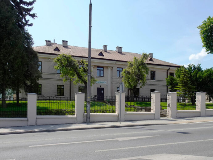 Two-story beige building behind a white fence with black iron railing, green lawn and trees, on a sunny day.