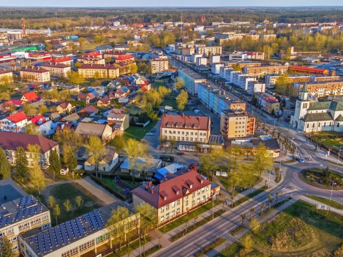 Aerial view of a small town with red-roofed houses, a church with a steeple, and a roundabout amid tree-lined streets and modern buildings.