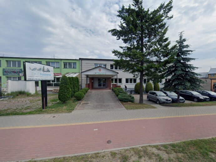 Public, two‑story light building with a central entrance, bordered by trimmed shrubs and a tall tree; a paved walkway leads to the door and a row of cars is parked to the right. A large billboard sits to the left of the entrance.
