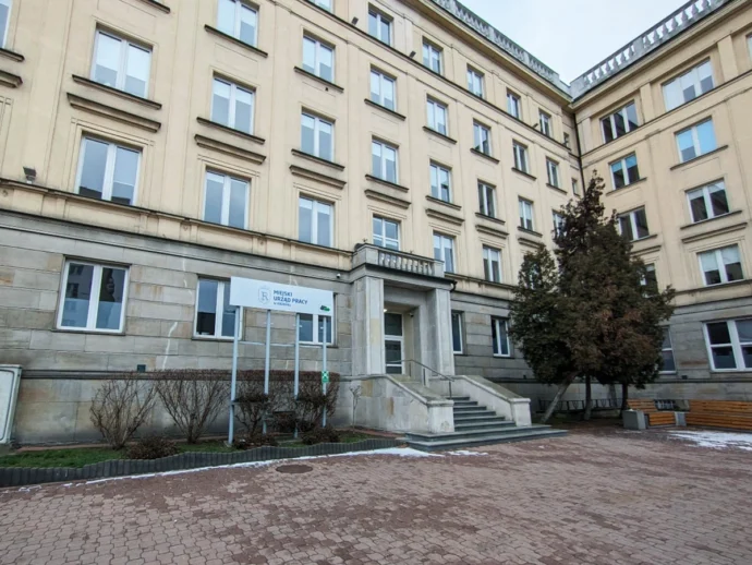 Facade of a beige, multi-story government building with a sign for Miejski Urząd Pracy at the entrance and a stone staircase.