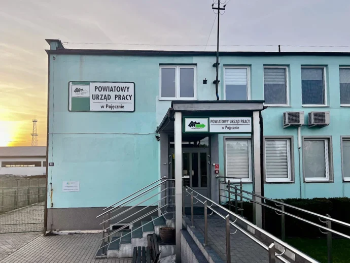Turquoise two-story office building with signs reading 'Powiatowy Urząd Pracy w Pajęcznie' and a ramp with metal handrails at the entrance.
