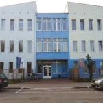 Three-story building with a blue central section and light-blue wings, glass entrance, bikes and cars parked outside.