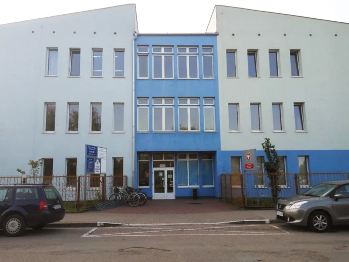Three-story building with a blue central section and light-blue wings, glass entrance, bikes and cars parked outside.