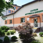 Building with green sign reading 'CENTRUM AKTYWIZACJI ZAWODOWEJ' and entrance, ramp, and bike rack outside amid shrubs and trees in a sunny day.