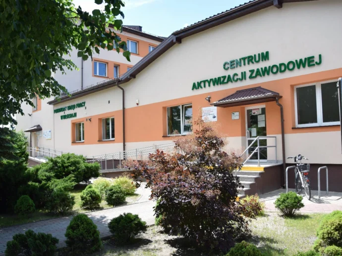 Building with green sign reading 'CENTRUM AKTYWIZACJI ZAWODOWEJ' and entrance, ramp, and bike rack outside amid shrubs and trees in a sunny day.
